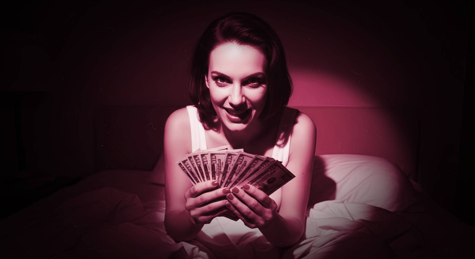 Smiling woman sitting on a bed under pink lighting, holding a fan of US dollar bills in both hands.