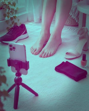 Adult creator photographing her feet anonymously with a smartphone, shoes, socks and nail polish nearby.