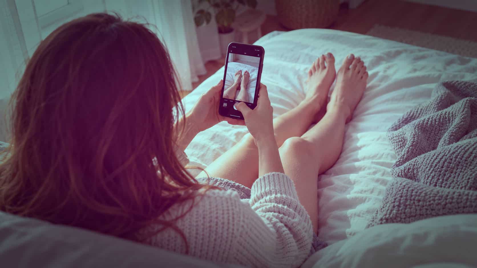 Taking Feet Photos at Home Without Showing Your Face Person sitting on a bed photographing their bare feet with a smartphone, viewed from behind with no face visible.